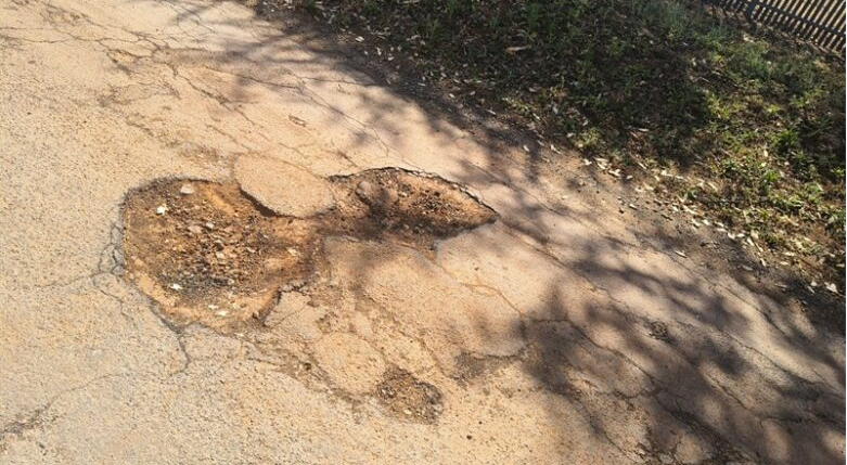  Wonderboom. Close-up of a massive pothole disrupting the cracked asphalt surface beside a chain-link fence. 